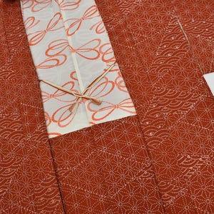 May include: A rust-colored haori jacket featuring a white geometric hemp leaf pattern and a white inner panel decorated with orange ribbon motifs. The garment includes two thin, light-colored ties at the center front for closure.