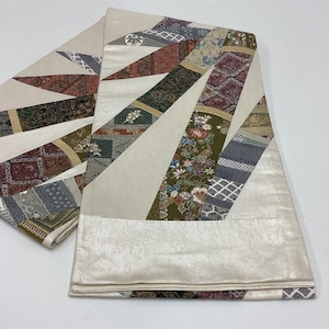 May include: A folded textile featuring a cream-coloured base with radiating diagonal bands. These bands display a variety of intricate patterns, including floral motifs, geometric shapes, and traditional Japanese-inspired designs in shades of green, red, brown, and blue.