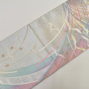 May include: A decorative textile with a light beige background and a colourful design. The design features vertical stripes in pink, blue, and green, along with stylized floral and leaf motifs. The textile is folded diagonally.