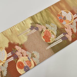 Può includere: Tessuto decorativo con sfumatura oro, marrone e verde. Il tessuto presenta motivi circolari ripetuti con motivi floreali e a ventaglio in rosso, arancione e blu. Il design include foglie stilizzate e motivi geometrici.