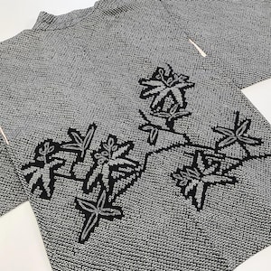 Puede incluir: Una parte superior estilo kimono con estampado en blanco y negro. La tela tiene un diseño texturizado y punteado. Bordados florales negros decoran la parte delantera y las mangas. La prenda está doblada, mostrando el escote y parte de las mangas.