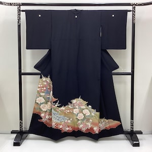 May include: A black kimono with a floral design on the hem, displayed on a black rack. The floral pattern includes pink and white flowers, with accents of green, red, and gold. The kimono is made of a dark fabric and has square sleeves.