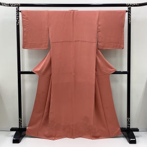 May include: A salmon-colored kimono hangs on a black display stand. The kimono features wide sleeves and a long, flowing design. The fabric appears smooth and has a subtle sheen. The stand has decorative elements on the top.