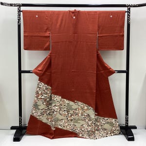 May include: A rust-colored kimono displayed on a black stand. The kimono features a textured pattern and a decorative lower border with a floral and geometric design. The sleeves are rectangular, and the garment is draped to showcase its form.