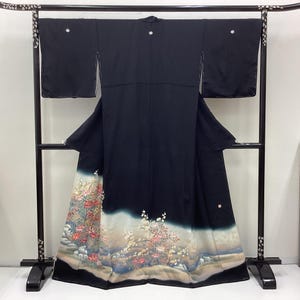 Puede incluir: Kimono negro con un diseño floral en el dobladillo, exhibido en un estante negro. El patrón floral presenta flores rosas, blancas y amarillas con hojas verdes sobre un fondo azul claro y beige. El kimono está hecho de una tela oscura.