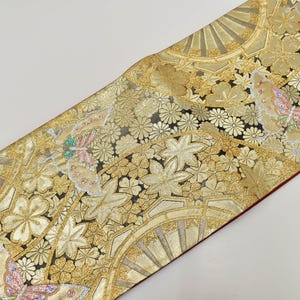 Può includere: Tessuto color oro con intricati motivi floreali e a farfalla. Il design presenta fiori, foglie e farfalle stilizzate in tonalità oro, rosa e verde acqua su sfondo nero. Il tessuto ha un bordo rosso.