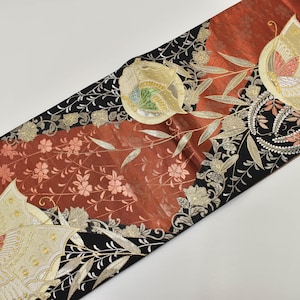 Può includere: Un runner da tavolo ornato con una ricca tavolozza di colori. Il runner presenta una sezione centrale in tessuto marrone rossiccio, bordata di nero e decorata con motivi floreali e a farfalla in oro e crema. Il design include ricami dettagliati.