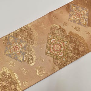 May include: A decorative textile with a repeating pattern of floral and geometric designs in shades of gold, brown, and red. The fabric has a subtle sheen, and the design is arranged diagonally across the surface.