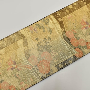 May include: A decorative textile featuring a gold background with detailed floral and bird motifs in peach, green, and cream. The fabric has a geometric border and a black edge. The textile is likely used for decorative purposes.