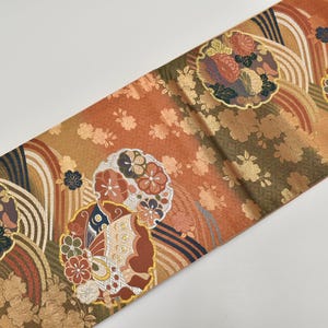 Può includere: Un tessuto ornato con un motivo ripetuto di medaglioni floreali e onde stilizzate. La tavolozza dei colori comprende sfumature di arancione, marrone, oro e blu navy. Il tessuto sembra essere seta o un materiale lussuoso simile, con una sottile lucentezza.