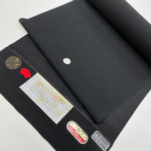 Pode incluir: Um rolo de tecido preto com uma superfície texturizada, parcialmente desenrolado para revelar um ponto branco e várias etiquetas anexadas. Os rótulos apresentam texto japonês e detalhes em vermelho, sugerindo um têxtil tradicional.