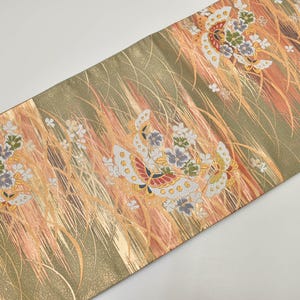 Può includere: Tessuto decorativo con un motivo ripetuto di farfalle ed elementi floreali. Il design presenta una tavolozza di colori oro, pesca e verde oliva. Il tessuto ha un aspetto strutturato, con una sottile lucentezza.