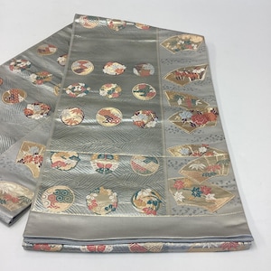 May include: A silver-toned fabric obi sash featuring a repeating pattern of circular medallions and fan-shaped motifs. The circular designs contain intricate floral and geometric patterns in red, gold, green, and white, while the fan motifs display detailed botanical illustrations against a shimmering, textured background.