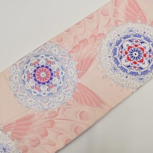 Può includere: Tessuto decorativo con un motivo ripetuto di disegni circolari a forma di mandala nei toni del blu, rosso e viola, su uno sfondo rosa pallido. Il design include elementi floreali e a forma di piuma stilizzati.