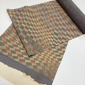 May include: A roll of dark fabric featuring a repeating geometric pattern of slanted rectangles in shades of orange, yellow, and green. The textile has a textured appearance with a dark gray border and a stamp on the unprinted white edge.