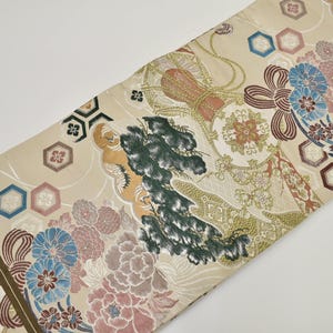 Può includere: Un tessuto beige con un motivo dettagliato di pini verde scuro, grappoli di fiori rosa e blu e nodi decorativi dorati. Il design include forme esagonali geometriche e un intricato lavoro di linee su tutta la superficie del tessuto.