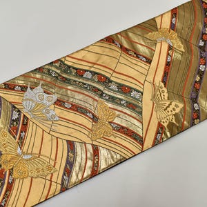 Può includere: Tessuto decorativo dorato e giallo con motivo a farfalle. Il tessuto presenta un motivo ripetuto di farfalle di varie dimensioni, con accenti dorati e argentati. Lo sfondo ha un motivo a righe con dettagli rossi, verdi e floreali.