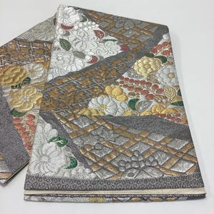 May include: A folded textile featuring a metallic silver and gold woven pattern with floral motifs in white, gold and green. The fabric displays geometric grid sections and textured dark grey panels, creating a complex, layered appearance.