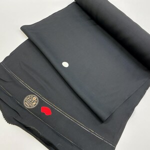 Pode incluir: Tecido preto, parcialmente enrolado, com um acento circular branco e costuras decorativas. Uma etiqueta com texto japonês e um acento vermelho é visível na parte dobrada. O tecido parece ser um têxtil.
