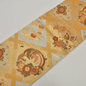 May include: A decorative textile with a repeating pattern of squares in gold, light blue, and tan. Each square features floral and bird designs in shades of orange, brown, and green. The textile is likely used for home decor.