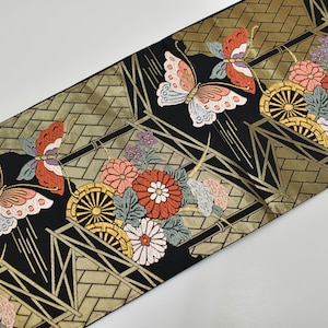 Può includere: Tessuto nero con un motivo geometrico dorato e ricami di farfalle e fiori. Le farfalle hanno ali rosse, rosa e bianche. I fiori sono rosa, rossi e bianchi. Il design include ruote stilizzate.