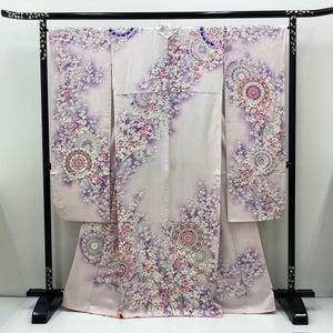 May include: A light pink kimono with a floral pattern. The kimono features a design of white and pink flowers with purple accents. The garment is displayed on a black metal rack.
