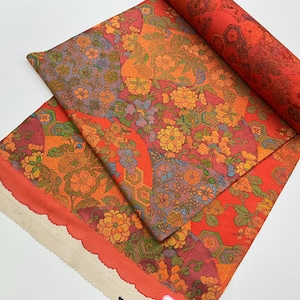 May include: A vibrant floral fabric with a repeating pattern of orange, yellow, and purple flowers on a red background. The fabric is partially rolled, revealing the detailed design. The edge of the fabric has a scalloped trim.