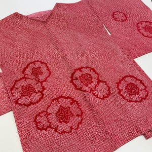 Puede incluir: Un kimono rojo con un patrón repetitivo de pequeños puntos blancos. Grandes diseños florales estilizados en un tono más oscuro de rojo se colocan sobre la tela. La prenda está doblada, mostrando el forro interior y los detalles del diseño.