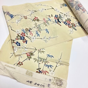 May include: A cream-colored fabric roll featuring a repeating pattern of plum blossoms and bamboo leaves in shades of red, blue, and white. The textile includes printed Japanese text and circular stamps along the bottom edge, indicating material composition.