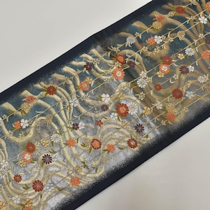 May include: A decorative textile with a dark blue border and a floral pattern. The design features orange, white, and purple flowers with gold and beige accents on a teal background. The fabric has a textured appearance.