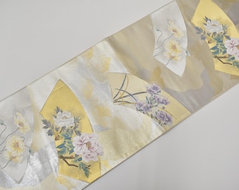 May include: A long, rectangular textile strip featuring a pale cream background with a subtle cloud pattern. The fabric is decorated with multiple fan-shaped motifs in gold and silver, each displaying detailed illustrations of blooming flowers.