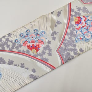 Può includere: Tessuto decorativo con un motivo ripetuto di forme a ventaglio floreali in rosso, blu e rosa su sfondo grigio chiaro. Il design include accenti floreali grigi e bordi rossi e grigi.