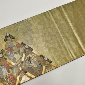 May include: Gold-toned decorative textile with a detailed floral and bird design. The textile features a repeating pattern of flowers and birds in shades of red, blue, and gold. The edges are trimmed with a black border. The textile is likely used as a table runner or decorative accent.
