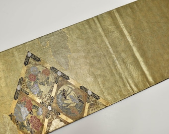 May include: Gold-toned decorative textile with a detailed floral and bird design. The textile features a repeating pattern of flowers and birds in shades of red, blue, and gold. The edges are trimmed with a black border. The textile is likely used as a table runner or decorative accent.