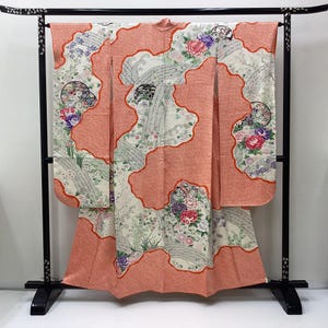 Puede incluir: Un kimono con una base de color crema y grandes paneles irregulares de color naranja coral. La tela está decorada con intrincados patrones florales, incluyendo peonías rosas, moradas y blancas, junto con motivos grises ondulados y diseños circulares.