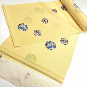 May include: A roll of pale yellow fabric featuring a repeating pattern of circular motifs. The circles contain intricate blue, red, and white geometric and floral designs. Some circles appear as faint, monochromatic green outlines on the fabric surface.