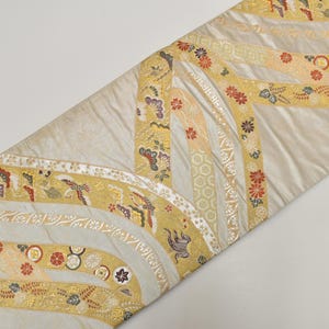 May include: A decorative textile with a cream background and gold, cream, and red floral and bird patterns. The design features curved bands with intricate details, creating a layered effect. The textile is likely used for home decor.