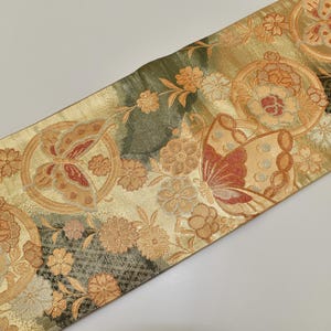 Può includere: Tessuto decorativo con un motivo ripetuto di farfalle e fiori nei toni dell'oro, dell'arancio e del crema. Il tessuto ha un aspetto strutturato e viene probabilmente utilizzato per scopi decorativi. Il design è intricato.