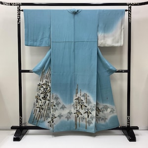 May include: A light blue kimono featuring a white gradient at the sleeves and hem. The lower section displays a painted pattern of dark brown and gold stylized floral motifs with vertical lines, suspended on a black wooden display stand.