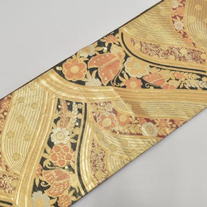 Può includere: Un tessuto decorativo con sfondo dorato e intricati motivi floreali nei toni dell'arancione, bianco e nero. Il design presenta fiori stilizzati e motivi a foglie, con un bordo nero.