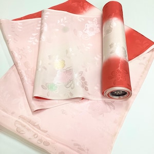 May include: A bolt of fabric featuring a gradient from pale pink to vibrant red, decorated with a tonal floral jacquard pattern. The textile is partially unrolled, revealing a smooth, lustrous surface with subtle, embossed flower motifs throughout.