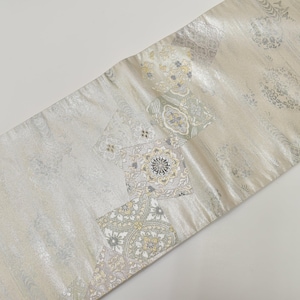 Puede incluir: Un camino de mesa rectangular y decorativo con un fondo beige claro brillante. El camino de mesa presenta un patrón repetido de formas cuadradas y rectangulares con diseños florales y geométricos en plata, gris y dorado.