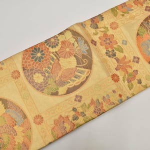 Può includere: Tessuto decorativo con un motivo ripetuto di motivi floreali e di uccelli in un design circolare. Il tessuto presenta uno sfondo color crema con accenti di arancione, blu e marrone. Il design è simmetrico e dettagliato.