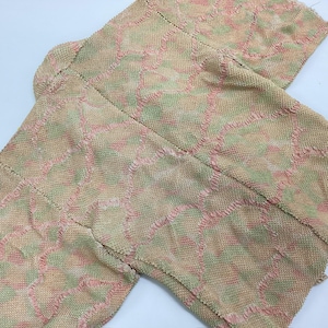 Puede incluir: Un kimono beige con un patrón texturizado en rosa y verde. La prenda está tendida, mostrando el intrincado diseño y los detalles de las costuras. La tela parece ligera y adecuada para superponer capas.