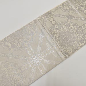 May include: A decorative fabric runner with a repeating pattern of silver and beige geometric shapes. The runner is made of a soft, textured material and is ideal for adding a touch of elegance to any table setting. The runner measures approximately 12 inches wide.