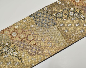 Pode incluir: Um têxtil decorativo com um design de patchwork. O tecido apresenta uma variedade de padrões em ouro, castanho, preto e branco. Os padrões incluem formas florais, geométricas e abstratas. O têxtil é provavelmente usado para decoração de casa ou moda.