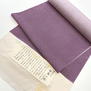 Op de afbeelding: Een opgerolde, paarse stof met een getextureerd oppervlak. Een crèmekleurig label met Japanse tekst is aan de stof bevestigd. De stof is effen van kleur met een subtiele glans. De presentatie suggereert een textielproduct.