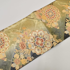 Puede incluir: Textil decorativo con fondo dorado y estampados florales en rojo, melocotón y amarillo. El diseño incluye flores y pájaros estilizados, con un borde negro en un lado. La tela tiene un patrón de cuadrícula geométrica.