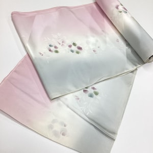May include: A roll of fabric featuring a soft gradient transition from pale pink to white and light grey. The textile is decorated with delicate, muted floral motifs in shades of purple, green, and grey, scattered across the surface.