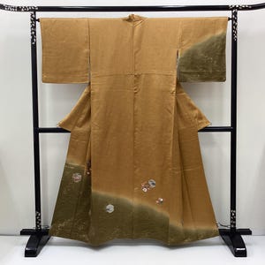 May include: A golden-brown kimono featuring wide sleeves and a gradient olive green hem. The garment is decorated with small, scattered floral motifs and hexagonal patterns near the bottom, displayed on a black wooden stand with mother-of-pearl inlay.
